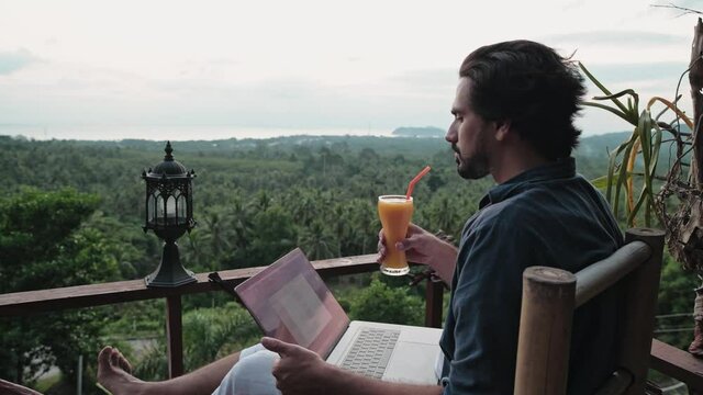 A young, handsome man with a beard works on a laptop in a cafe overlooking an incredible sunset.