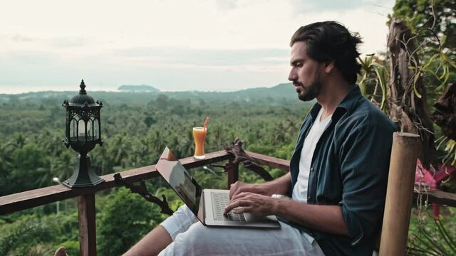 A young, handsome man with a beard works on a laptop in a cafe overlooking an incredible sunset.
