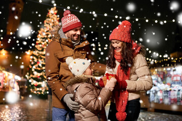 Fototapeta na wymiar family, winter holidays and celebration concept - happy mother, father and little daughter with gift at christmas market on town hall square in tallinn, estonia over snow