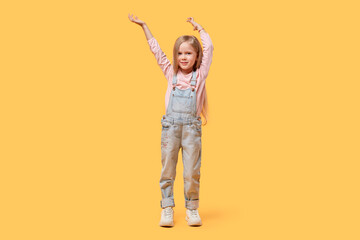 Portrait of a little blonde girl happily raised her hands up to her full height. He smiles and looks at the camera. Yellow isolated background.