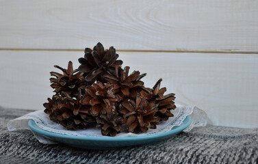 Pine cones of dark brown and golden brown on a white linen scarf with lace lying on a pale turquoise plate against a background of white boards and a gray sweater. New Year. Christmas. Winter. Holiday