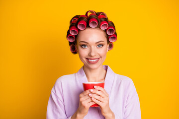 Close-up portrait of charming cheerful woman wearing curlers drinking warm beverage isolated over vivid yellow color background
