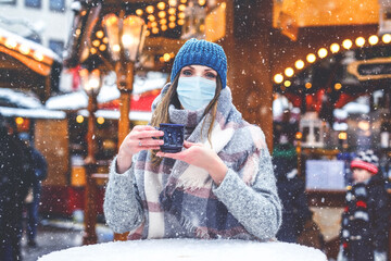 Woman with medical mask drinking hot punch, mulled wine on German Christmas market. People with...