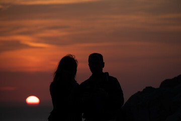 Fototapeta na wymiar Young couple in love admiring a beautiful red and orange sunset in a famous sunset point in Sardinia, Italy. Romantic moment for the two, framed by the sun an the scenic sky.