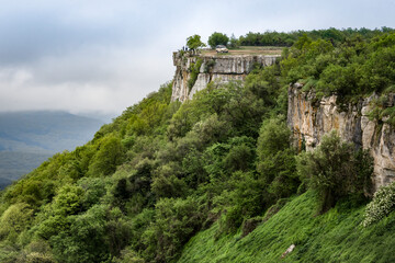 Fototapeta premium BAKHCHISARAI, RUSSIA - 7 MAY, 2019: The rock above the mountain valley with a car on top in the fog on the background of a stormy sky near the town of Bakhchisarai in the Crimean Mountains