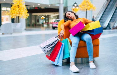 Tired and stressed young woman is lying and resting in the chair  with many shopping bags in the...