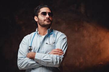 Self confident and brutal doctor with sunglasses in white labcoat poses with crossed arms in dark...