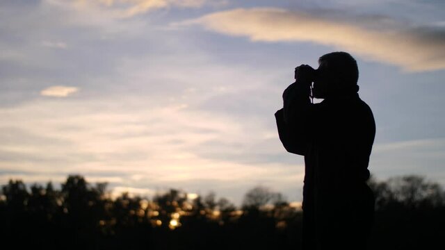 Silhouette of man using binoculars during beautiful sunset and epic sky. Impressive slow-motion scene of birdwatching and exercising hobby. Beautiful nature background during fall. Happy lifestyle.