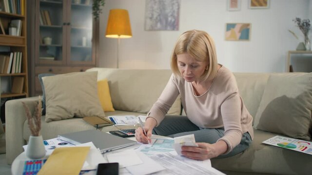 Medium footage of middle aged caucasian female sitting at table with piles of documents and payments filling out tax declaration form smiling
