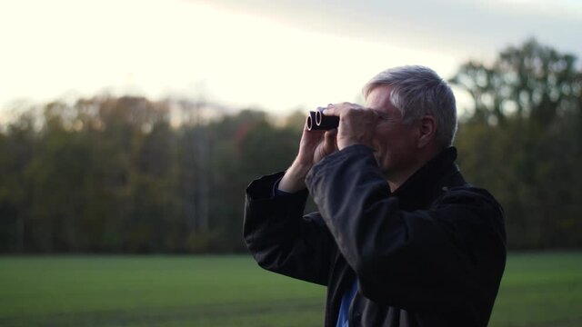 Attractive middle-aged white man using binoculars in a forest in late autumn. Handsome guy watches birds outdoors and enjoys his hobby. Beautiful nature background during sunset. Happy lifestyle.