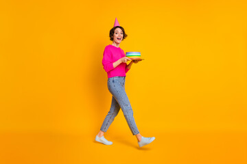 Full body photo of youngster lady hold birthday cake walk wear pullover cone hat jeans shoes isolated yellow color background