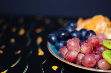 Fruits, grapes, tangerines, bananas, kiwi in a blue porcelain plate. black wooden floor, fruit plate