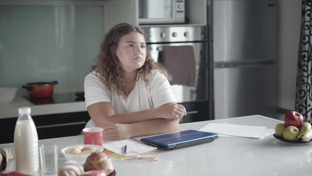 Lonely sad chubby woman sitting in kitchen at home and sighing. Portrait of upset young Caucasian overweight lady spending morning alone at home. Loneliness concept.