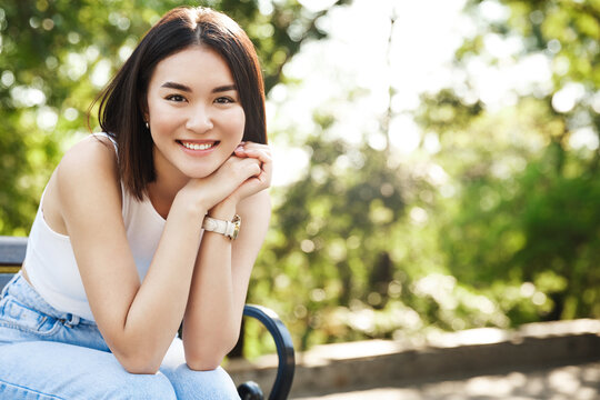 Beautiful asian woman sitting on bench and smiling. Modern girl resting in park on sunny summer day, looking happy