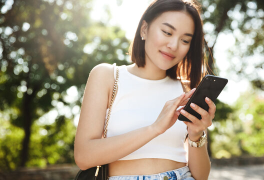 Asian young woman standing in park and reading message on mobile phone. Girl smiling while looking at her photos on smartphone