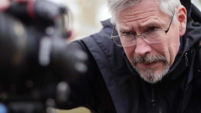 Mature photographer taking pictures in nature with a professional DSLR camera. Male Caucasian looks at the camera to check photos or change settings. Outdoors. Using a tripod, camera stand.