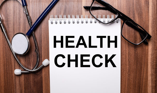 HEALTH CHECK is written on white paper on a wooden background near a stethoscope and black-framed glasses. Medical concept