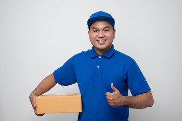 Young smiling asian delivery man in blue uniform holding box parcel cardboard and showing thumbs up on isolated white background.