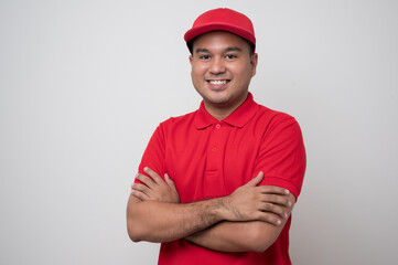 Smiling young handsome asian delivery man in red uniform standing with arm crossed on isolated white background.