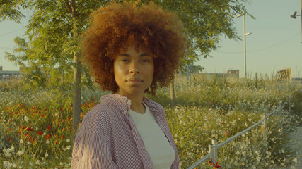 Portrait of mixed race black woman in the park full of flowers with huge afro hair in contour sunlight watching to the camera. Cinema film colors - 390097153