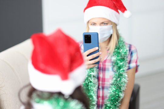 Woman in protective medical mask and santa claus hat sits on couch and takes pictures of her girlfriend on smartphone. Christmas with coronavirus pandemic concept