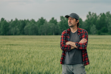 Naklejka na ściany i meble Wheat farmer agronomist standing in cultivated cereal crops agricultural field with arms crossed and looking into distance with uncertain facial expression