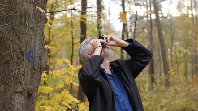 Mature middle-aged white man taking pictures in a scenic forest in late autumn. Handsome male photographer travels outdoors with a small camera. Beautiful nature background during fall. Happy tourist.