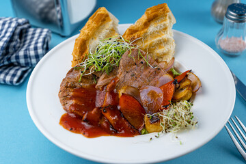 Warm salad with beef, vegetables, croutons and microgreen in a white plate on the restaurant table. Meat food in a bowl. Chop, medallions, cutlets. Blue background, close-up.