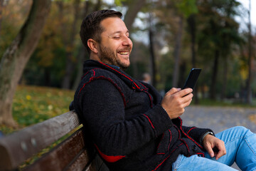 Man smiling and sitting on the bench in the park at autumn with smartphone in his hand