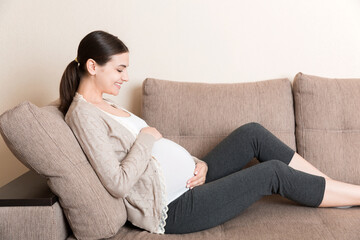 photo of cute caucasian future mom lying in light living room on sofa and holding hands on tummy