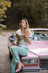 young woman looking away while sitting on hood of retro cabriolet and holding book on blurred background