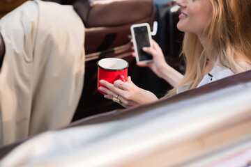 partial view of young woman holding smartphone with blank screen and cup of coffee on blurred foreground
