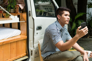 Man sitting on chair using smartphone near RV car in camping site.