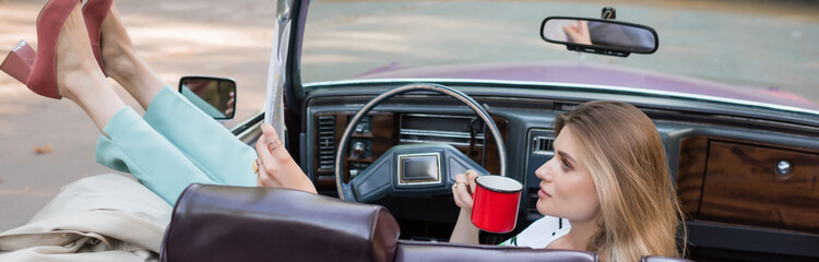 young woman holding cup of coffee and looking at map while sitting in cabriolet with legs raised on...