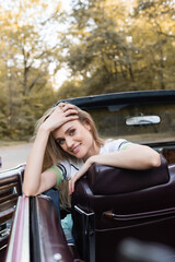 cheerful woman sitting in vintage cabriolet, touching hair and looking at camera, blurred foreground