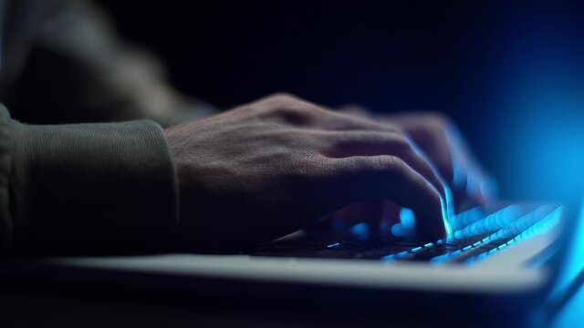 Extreme close-up hands of unrecognizable hacker programmer working typing on keyboard laptop computer at dark room. Freelancer using laptop working from home, soft blue color. Shooing in slow motion.