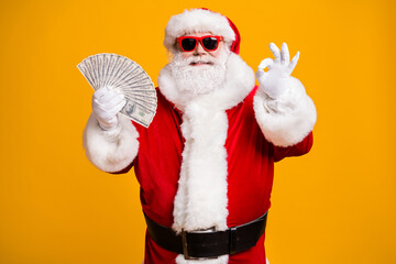Portrait of his he nice attractive cheerful fat overweight bearded Santa holding in hand cash budget showing ok-sign good job advert isolated bright vivid shine vibrant yellow color background