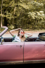 Fototapeta na wymiar smiling woman in sunglasses touching hair and looking away while sitting in vintage cabriolet