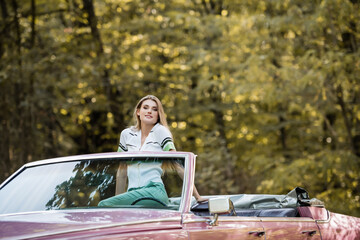 young stylish woman looking at camera while posing in vintage convertible car