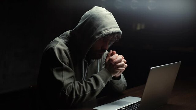 Thoughtful Caucasian hacker man looking at laptop screen and working on computer thinking solving problem. Side view of dangerous concentrated young guy hacking online web site at dark room.