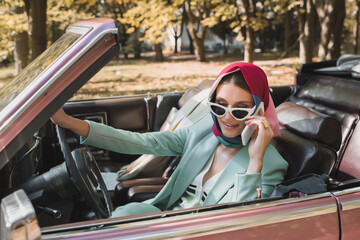 Positive woman in sunglasses talking on smartphone in roofless auto on blurred foreground