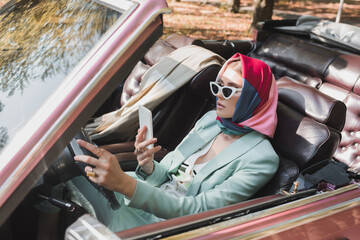 Elegant woman using smartphone while driving roofless car on blurred foreground