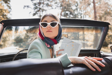 Stylish woman in sunglasses holding map in retro car on blurred foreground during trip