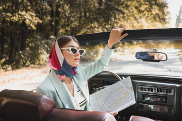 Elegant woman holding map while sitting in roofless car on road on blurred foreground