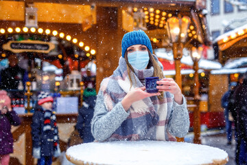 Woman with medical mask drinking hot punch, mulled wine on German Christmas market. People with...
