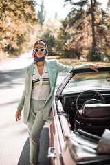Elegant woman in sunglasses standing near vintage auto during trip on blurred foreground