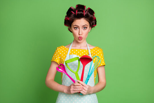 Photo of cute girl hold kitchen equipment whisk spoon fork spatula potato masher make her lips pouted wear yellow dotted t-shirt isolated over green color background
