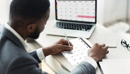 Black Businessman Using Calendar Program On Laptop And Writing Notes To Planner