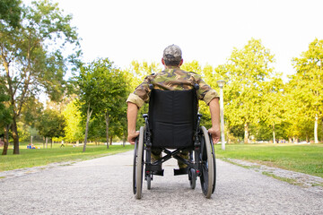 Single disabled retired military man in wheelchair moving down footpath in city park. Back view....