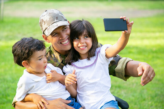 Two cheerful kids sitting on dads lap and taking selfie on cell. Disabled military man walking with kids in park. Veteran of war or disability concept